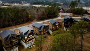 A cluster of log cabin-style houses with multiple stories and balconies, surrounded by trees and a distant town. - Lux Home-2 Master Suites-Heated Pool-Dollywood 5mi (Pigeon Forge)