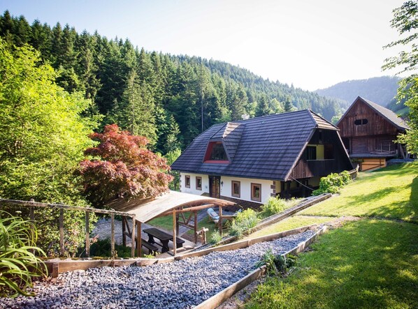 A rustic wooden house with a sloped roof, surrounded by a garden and a gravel path.