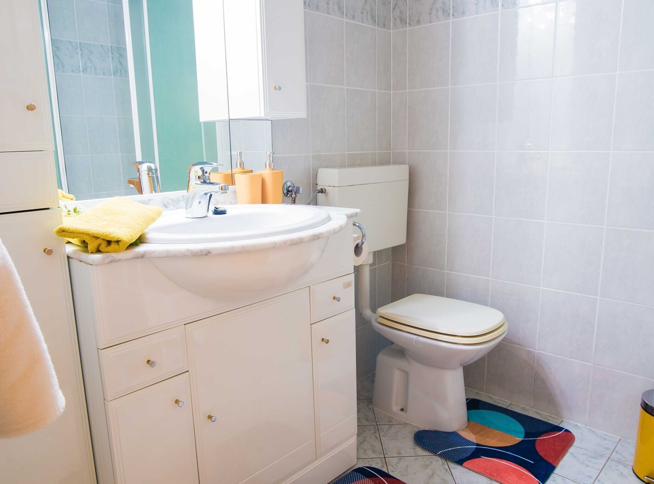A bathroom with a white sink, toilet, and cabinets. There is a yellow towel on the sink and a colorful mat on the floor.