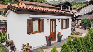 Un edificio bianco con tetto in tegole, persiane in legno e una porta con un’apertura superiore. Vasi con fiori rossi adornano il terreno di fronte all'edificio.