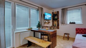 A cozy room with a wooden bench, a TV on a wooden shelf, a bed, and large windows with blinds.