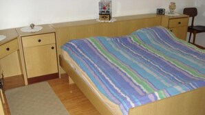 Uma cama de solteiro com um edredom colorido listrado, uma mesa de cabeceira de madeira com um rádio e uma pequena mesa com um vaso e um relógio.
