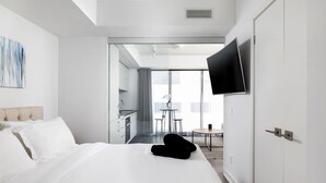 A modern hotel room with a large bed, a flat-screen TV mounted on the wall, a small dining area, and a view of the city.