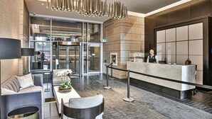 A modern hotel lobby with a reception desk, seating area, and a glass elevator.