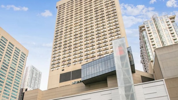 Un bâtiment moderne à la façade structurée en forme de grille, entouré d’immeubles de grande hauteur.