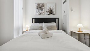 Un lit bien fait, avec du linge de lit blanc et une serviette enroulée, deux tableaux représentant des fleurs accrochés au mur, et des lampes de chevet posées sur les tables de nuit.