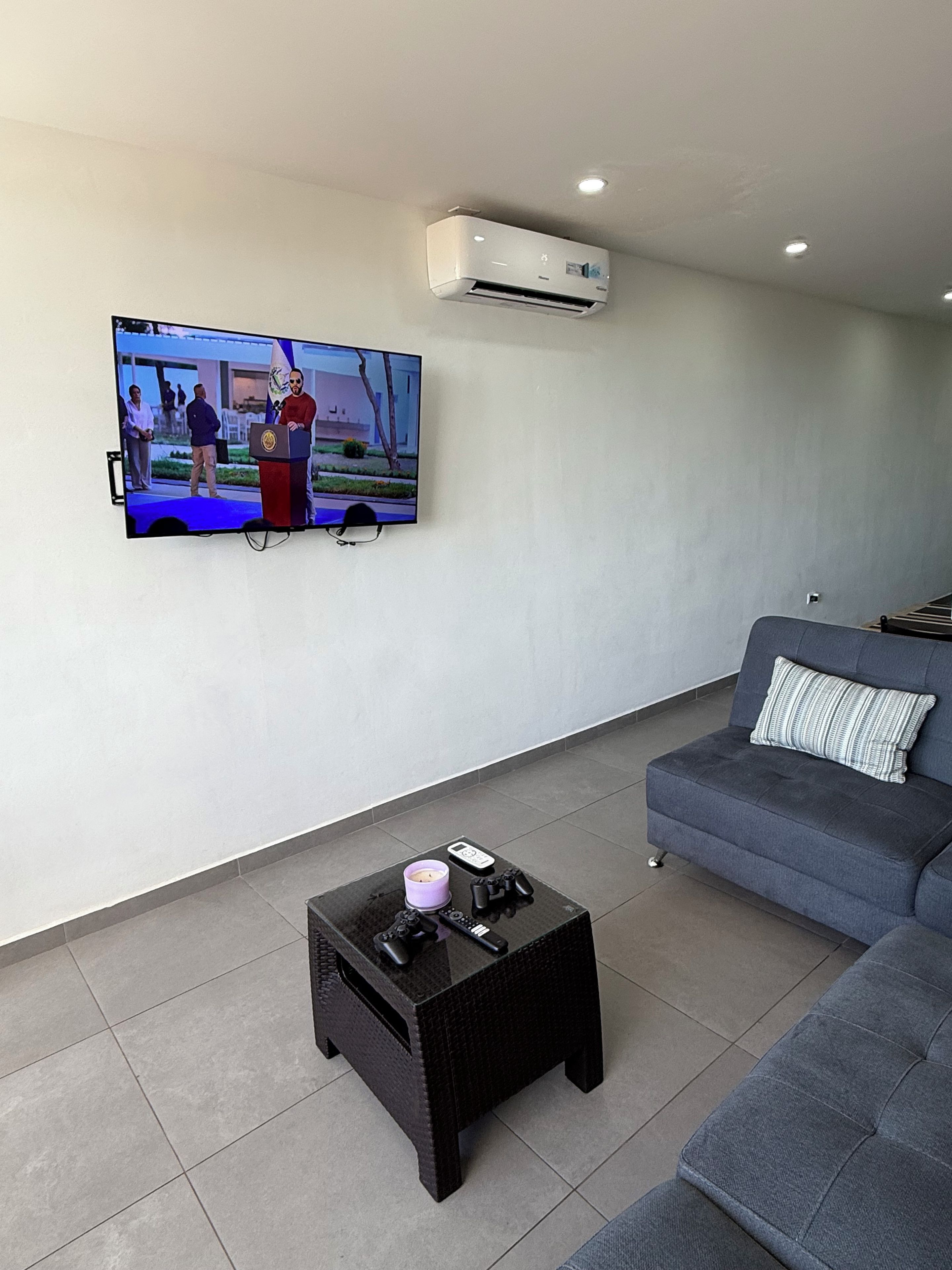 Una sala de estar moderna con un televisor de pantalla plana montado en la pared, una mesa de centro negra con un control remoto, y un sofá gris con un cojín a rayas.