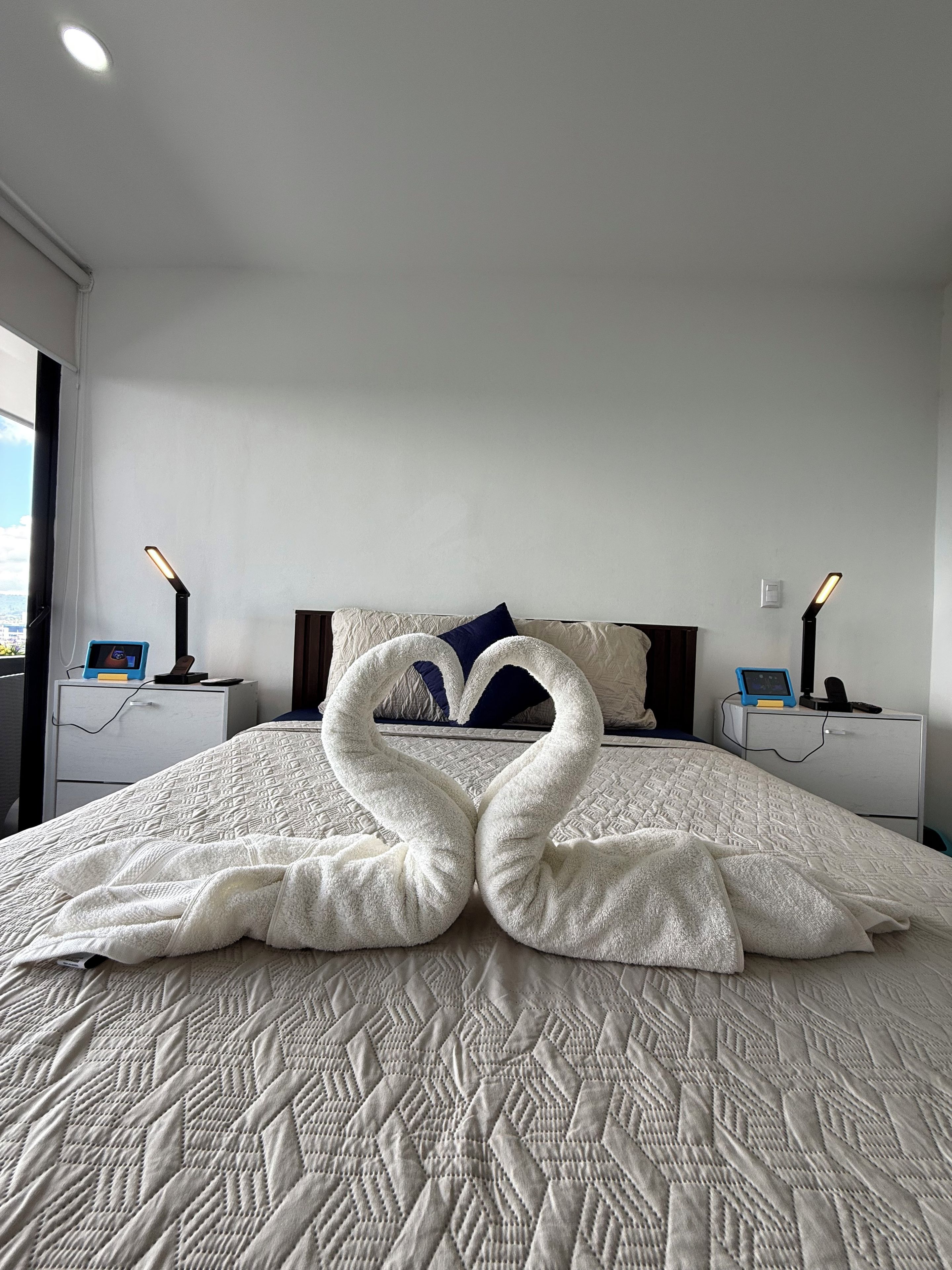 A hotel room with a bed, two bedside tables with lamps, a digital clock, and a heart-shaped towel decoration on the bed.