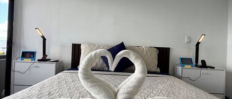 A hotel room with a bed, two bedside tables with lamps, a digital clock, and a heart-shaped towel decoration on the bed.