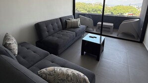 Ein modernes Wohnzimmer mit einem dunkelgrauen Sofa, gemusterten Kissen, einem kleinen schwarzen Couchtisch und einem großen Fenster mit Blick auf Grünflächen und Gebäude.