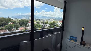 A balcony with a view of a cityscape and mountains, a white desk with a lamp, and a chair.