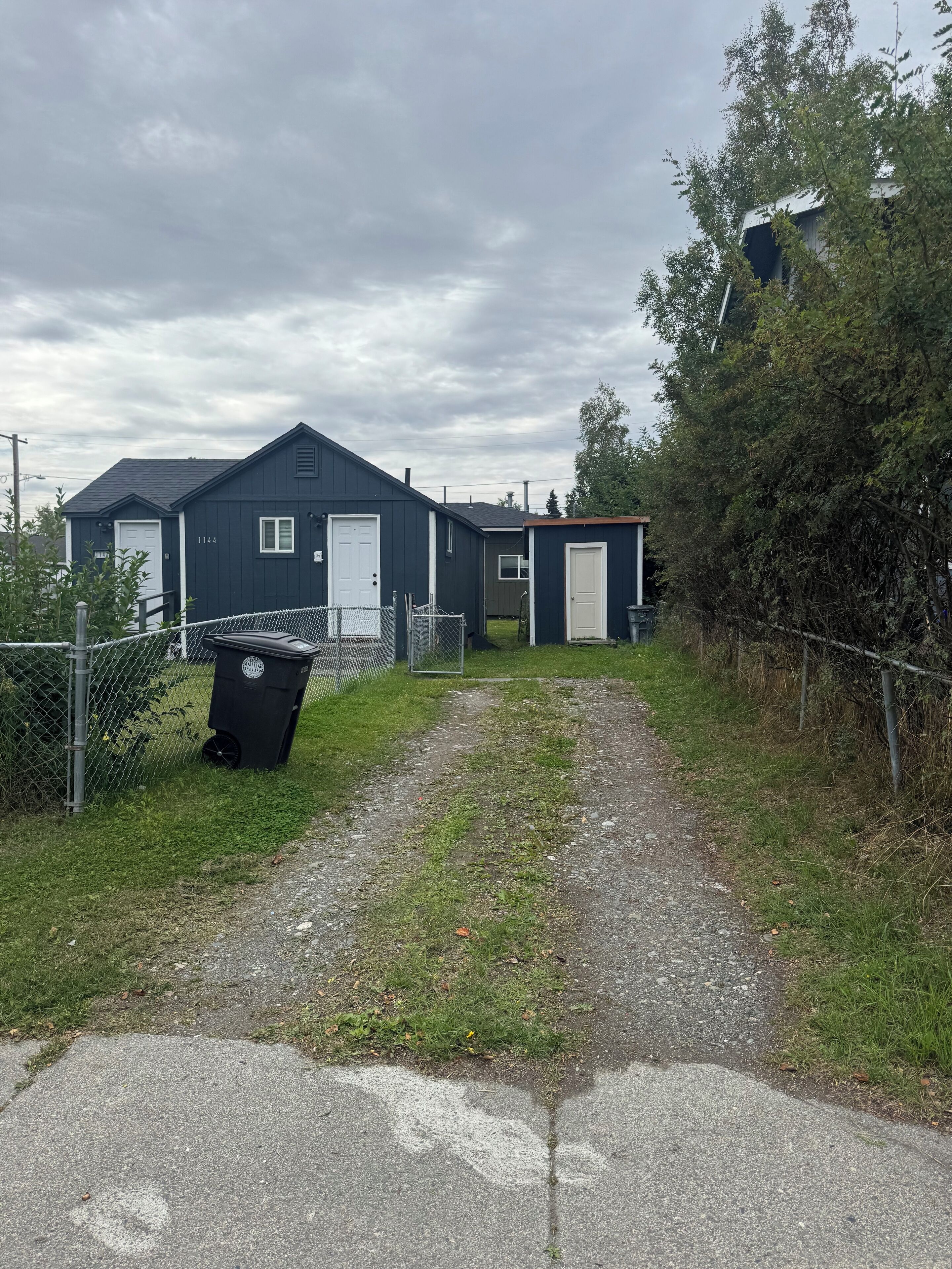 A pathway leading to a dark blue building with a white door, a smaller attached structure, and a black trash bin.
