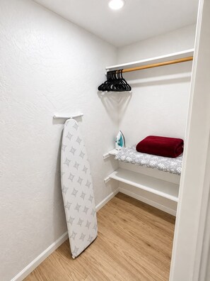 A small, well-organized closet with a hanging rack, a shelf with a red towel, and a white ironing board. - Cozy house in charming Anchorage with WiFi access (Anchorage)