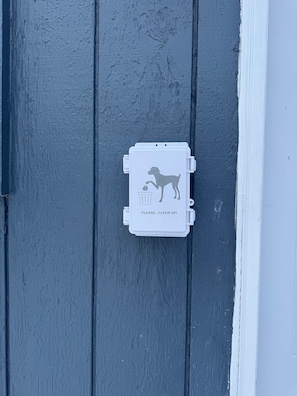 Une poubelle blanche avec un chien et un pictogramme représentant une poubelle, et le texte « VEUILLEZ… NETTOYER ! » sur un mur gris.