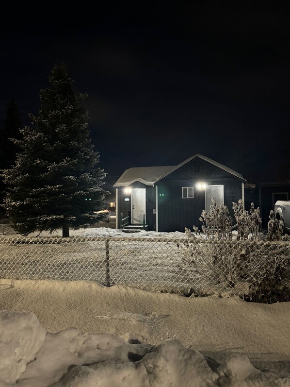 Une maison avec une pelouse recouverte de neige, une clôture en bois et un arbre également recouvert de neige.
