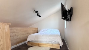 A small, sloped bedroom with a wooden bed frame, a white mattress, and a fluffy white rug. There is a wall-mounted TV and a small window providing natural light. - Glacier Alpine Lodges (East Glacier Park)