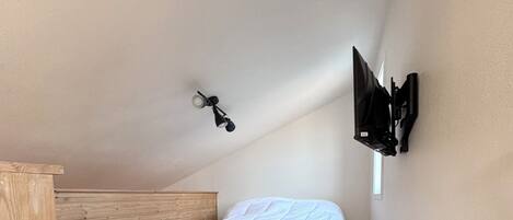 Un dormitorio pequeño con techo inclinado, estructura de cama de madera, colchón blanco y alfombra mullida. Hay un televisor montado en la pared y una ventana que deja entrar luz natural.