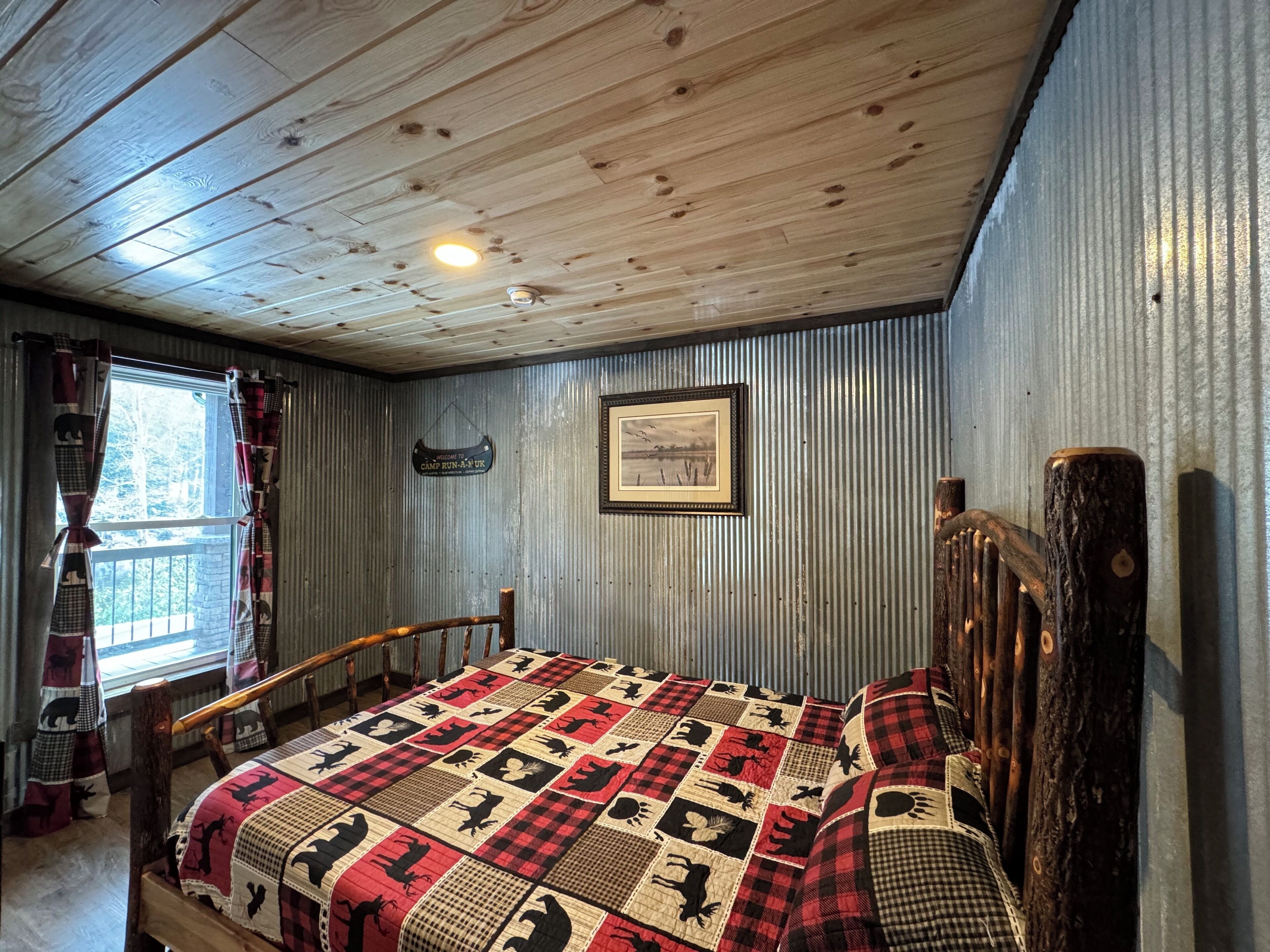 A cabin-style room with a wooden ceiling, a bed with a checkered quilt, a window with curtains, a framed picture on the wall, and a wooden headboard.