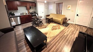 A living room with a sofa, a coffee table, a dining table with stools, a kitchen area with cabinets and appliances, and a door with a handle. - Charming apartment on Main Street  (Lake Placid)