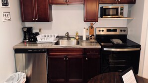 A compact kitchen with dark wooden cabinets, a stainless steel dishwasher, a microwave, a stove, and a coffee maker. - Charming apartment on Main Street  (Lake Placid)