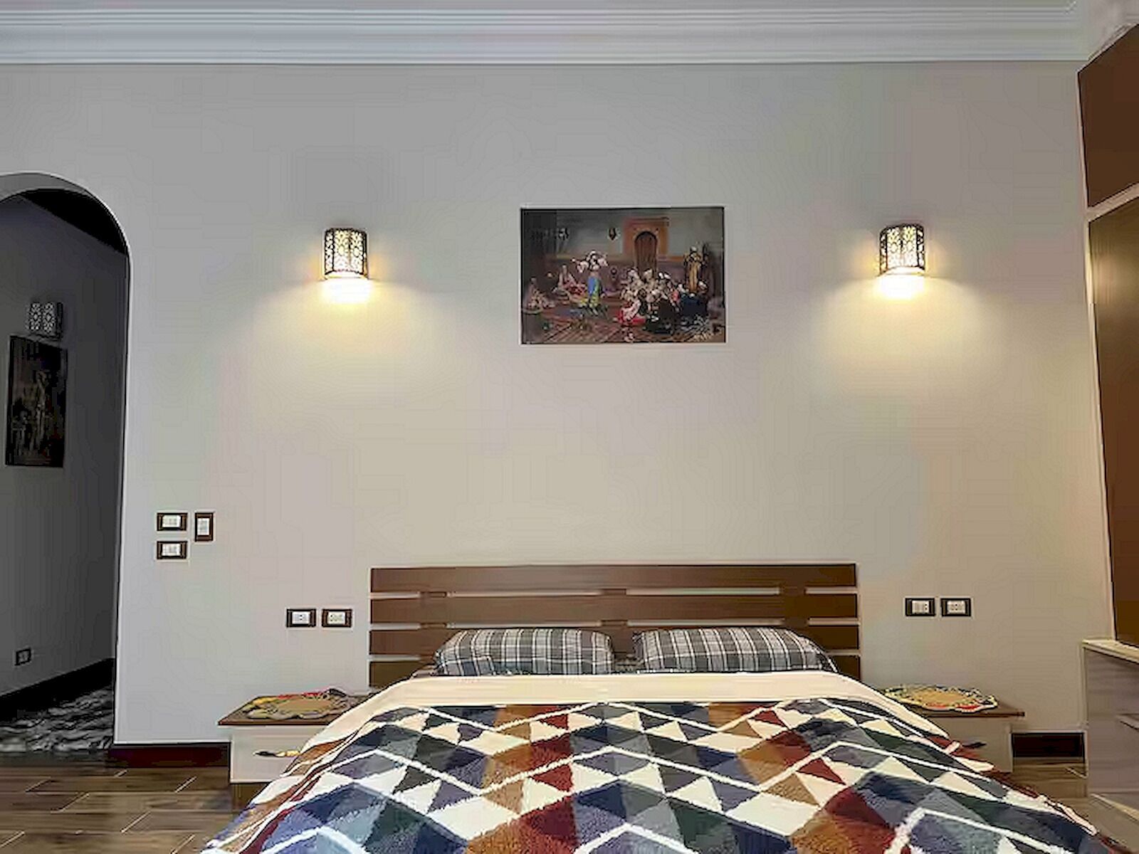 A bedroom with a large bed, two bedside tables, a painting on the wall, and two wall-mounted lamps.