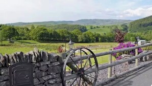 A stone wall with a mailbox and a wheelbarrow, overlooking a green landscape with hills.