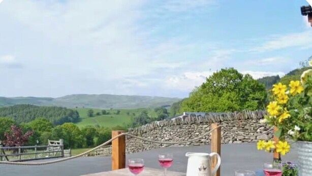 A wooden table with a pitcher and glasses of pink liquid, set against a scenic backdrop of hills and a stone wall.