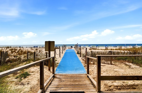 A wooden boardwalk leading to a beach with a blue mat, surrounded by sand dunes and wooden fences.