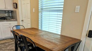 A dining area with a wooden table and black chairs, a window with blinds, and a kitchen with white cabinets. - Rock Scene Arcade, Retro Rock & Arcade games. (Houston)