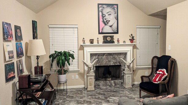 A living room with a fireplace, a mounted picture, a potted plant, and a chair with a pillow. - Rock Scene Arcade, Retro Rock & Arcade games. (Houston)