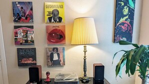 A room with a wall decorated with various album covers, a table with a lamp and speakers, and a small figurine. - Rock Scene Arcade, Retro Rock & Arcade games. (Houston)