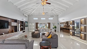 A spacious living area with a high ceiling, built-in shelving, a large flat-screen TV, and a dining area with a kitchen in the background.