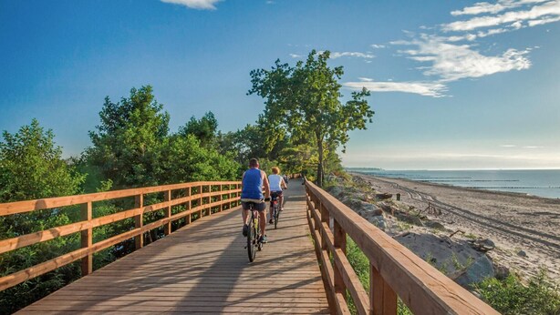 A wooden boardwalk with cyclists, surrounded by greenery and leading to a beach. - Amazing apartment in Ustronie Morskie (Ustronie Morskie)