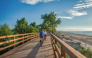 A wooden boardwalk with cyclists, surrounded by greenery and leading to a beach.