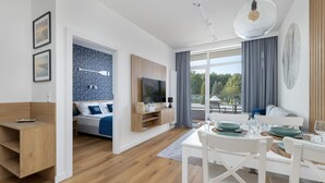 A modern, well-lit room with a wooden floor, a bed with blue bedding, a TV, a dining table set for four, and a large window with curtains. - Amazing apartment in Ustronie Morskie (Ustronie Morskie)