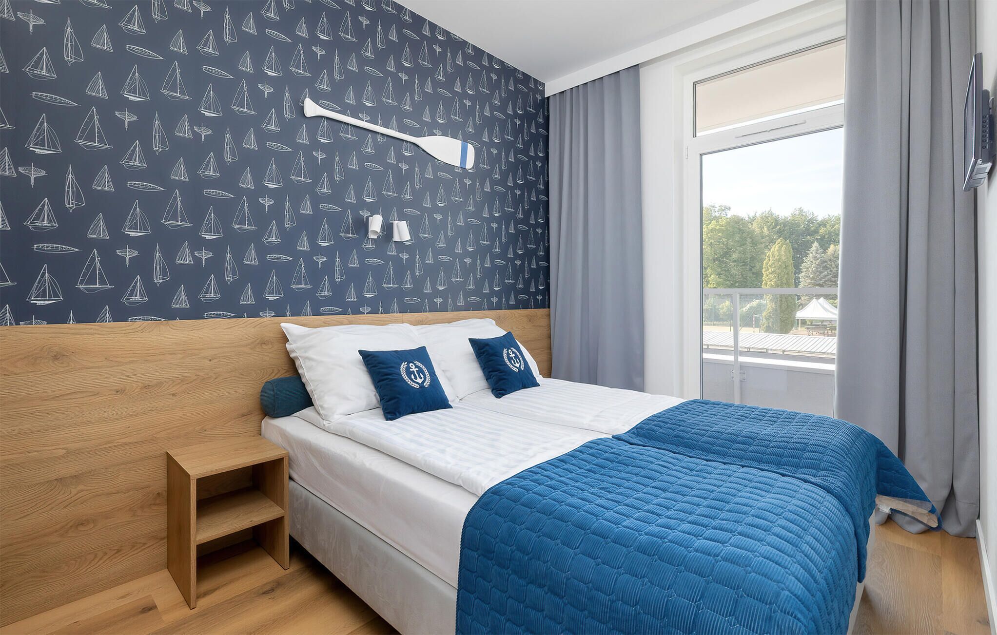 Une chambre à coucher avec un lit, une table de chevet, une fenêtre avec des rideaux et un mur orné d’un motif représentant un voilier.
