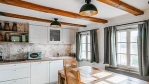 Une cuisine avec des meubles blancs, une table en bois et un grille-pain vintage. On y trouve deux lampes suspendues et une fenêtre avec des rideaux.