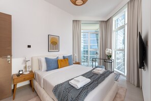 A modern hotel room with a large bed, a bedside table with a lamp, a small table with a plant, and a view of the city through large windows. - Vibrant 2BR, Miami Vibes at Business Bay (Dubai)
