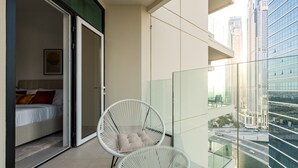A balcony with a glass railing, a bed with pillows, and a white wicker chair. - Vibrant 2BR, Miami Vibes at Business Bay (Dubai)