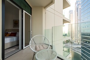 A balcony with a glass railing, a bed with pillows, and a white wicker chair. - Vibrant 2BR, Miami Vibes at Business Bay (Dubai)