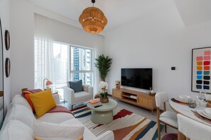 A modern living room with a large window, a flat-screen TV, a dining area, and a sofa with cushions.