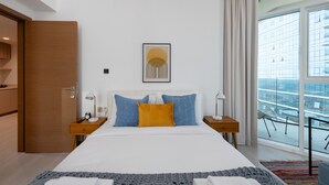 A modern hotel room with a large bed, two bedside tables, a window with a city view, and a wooden door. - Vibrant 2BR, Miami Vibes at Business Bay (Dubai)
