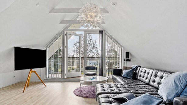 Un dormitorio tipo loft moderno con un tragaluz grande, un sofá de cuero negro, una mesa de centro redonda y un televisor de pantalla plana sobre un soporte de trípode.