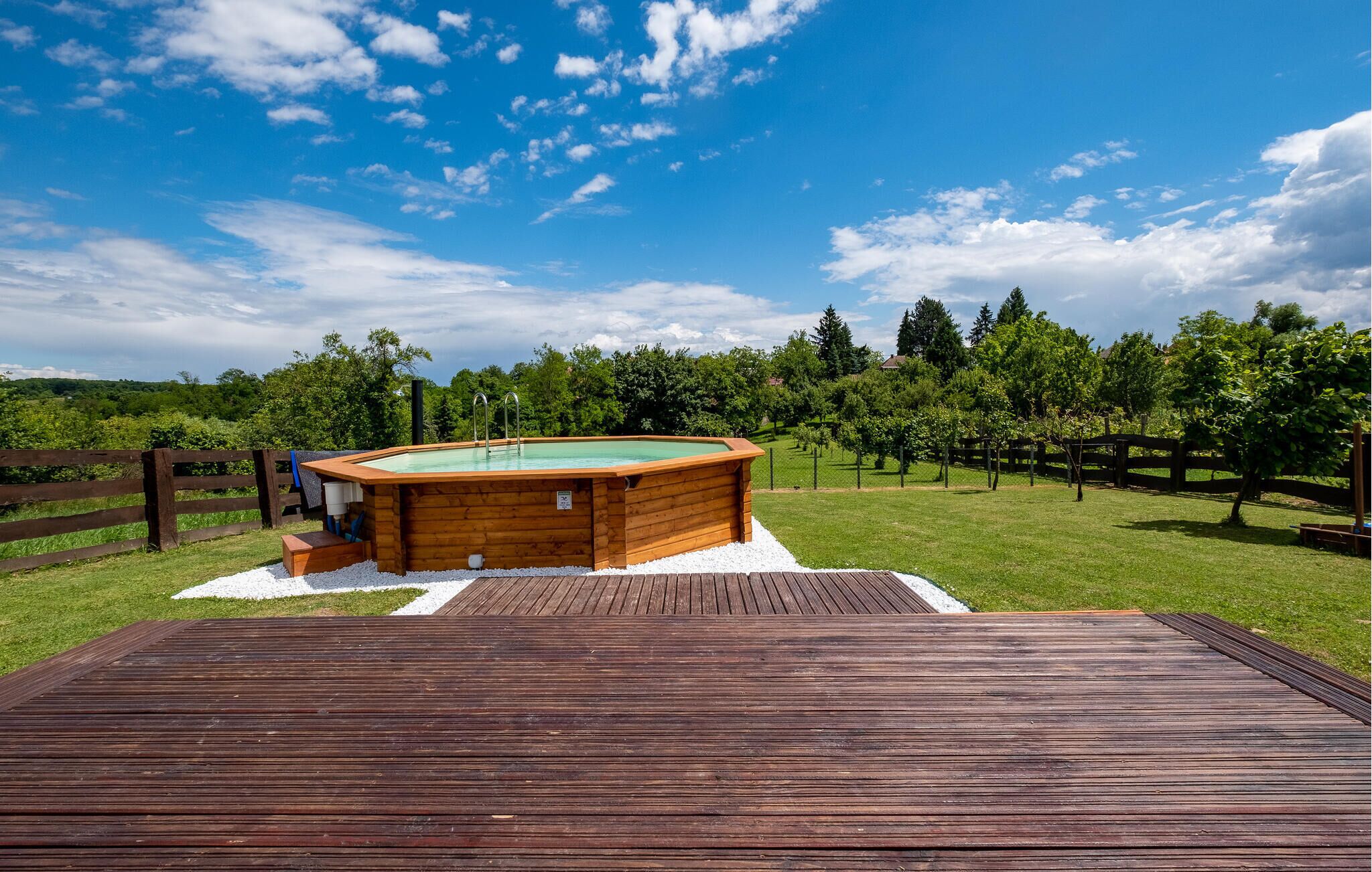 Eine Holzterrasse führt zu einem Whirlpool in einer grünen Umgebung mit Bäumen im Hintergrund.