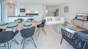 A modern kitchen with white cabinets, a dining area with a wooden table and black chairs, a white sofa, and a television on a wooden stand. - Cozy apartment in Oostende with WiFi (Oostende)