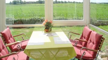A conservatory with a table set for four, surrounded by red cushioned chairs, overlooking a green field.