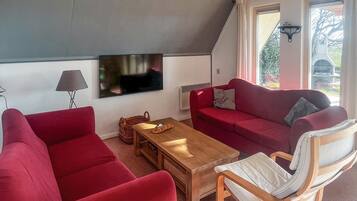 A cozy living room with a red sofa, a wooden coffee table, and a white armchair. A flat-screen TV is mounted on the wall, and there is a window with a view of greenery outside.