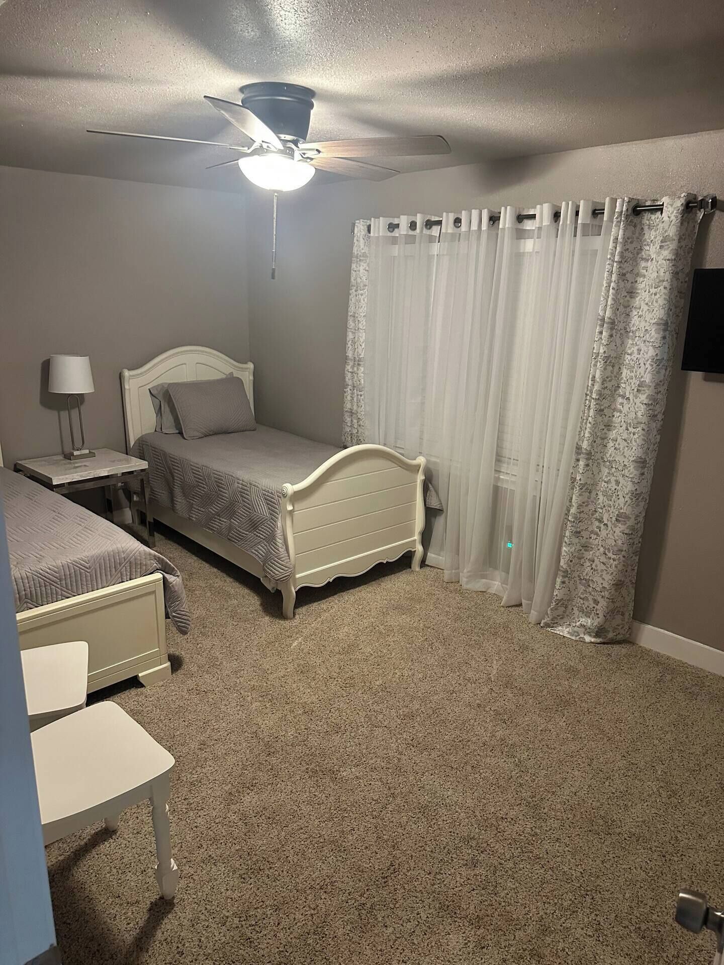 A bedroom with a bed, a nightstand, a lamp, a ceiling fan, and a window with curtains.