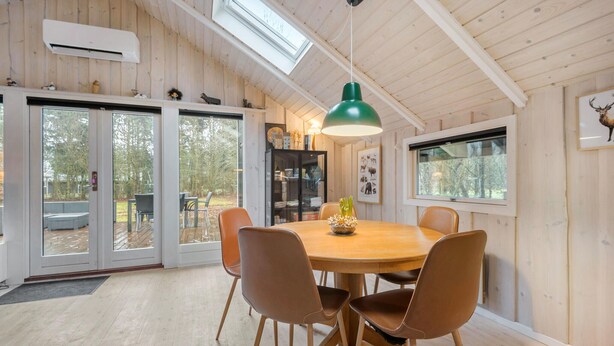 A wooden interior with a round table and six chairs, a green pendant light, and a view of the outdoors through large glass doors. - Gorgeous home in Oksbøl with sauna (Oksbøl)