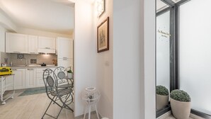 Eine moderne Küche mit weißen Schränken, ein Essbereich mit einem Metallstuhl und Tisch, und ein kleiner weißer Hocker. In der Nähe einer Glastür befinden sich zwei Blumentöpfe mit Spiegelung.